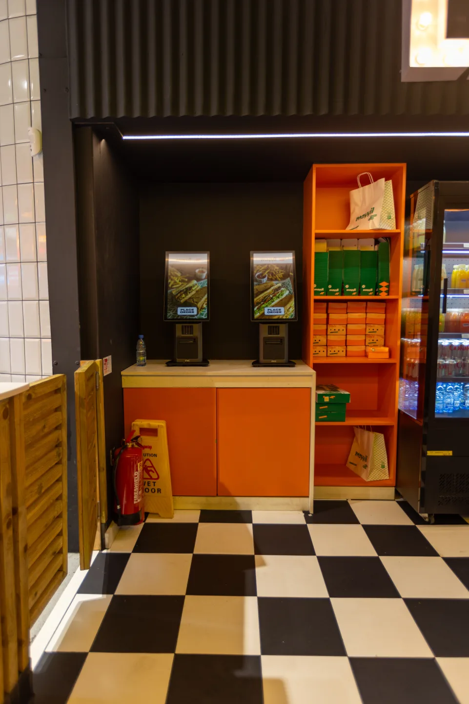 Self-order kiosk station with two terminals on a spray-painted orange cabinet base — Mayyil, Notting Hill