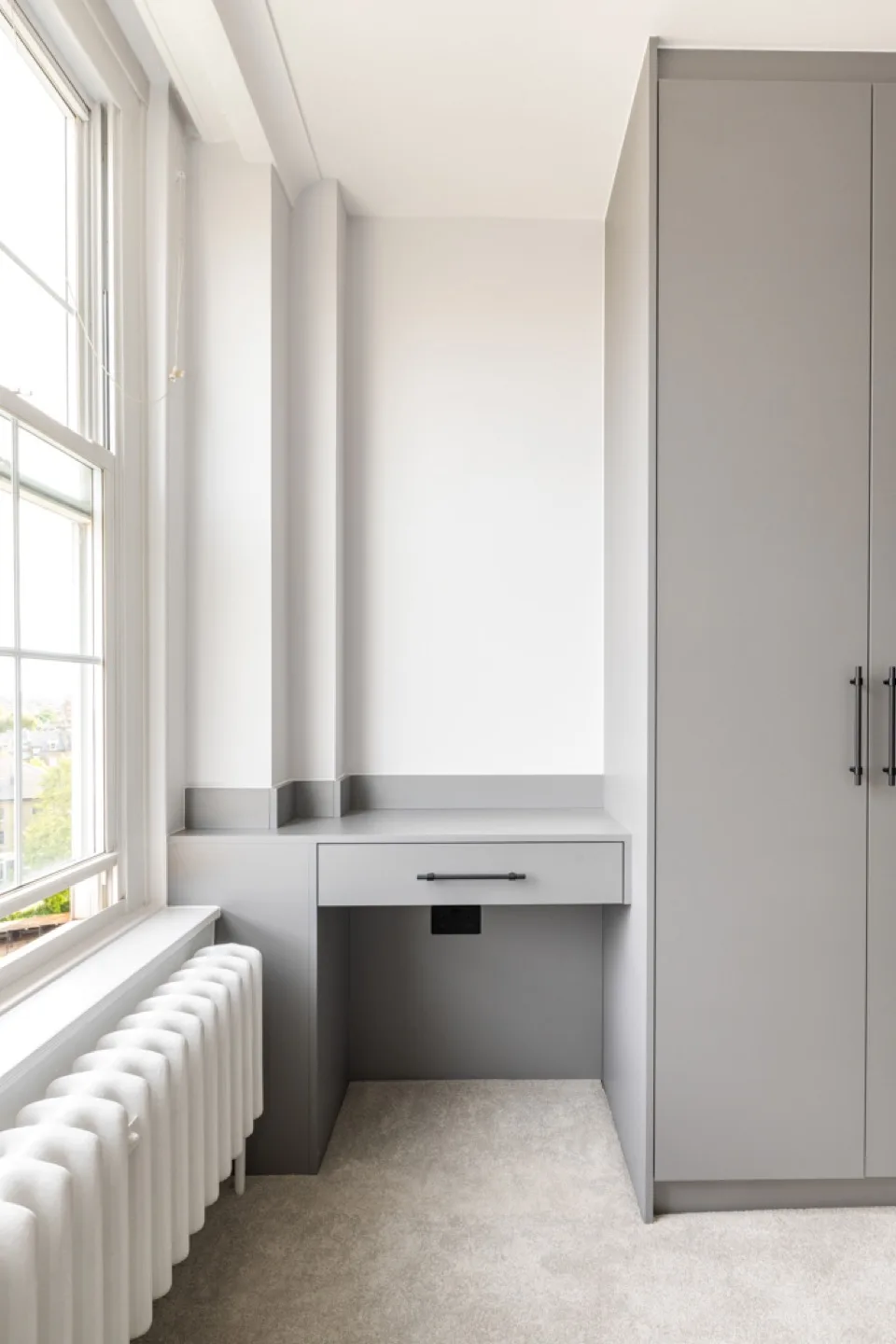 Compact grey floating desk with a drawer fitted beside a tall grey wardrobe by the window