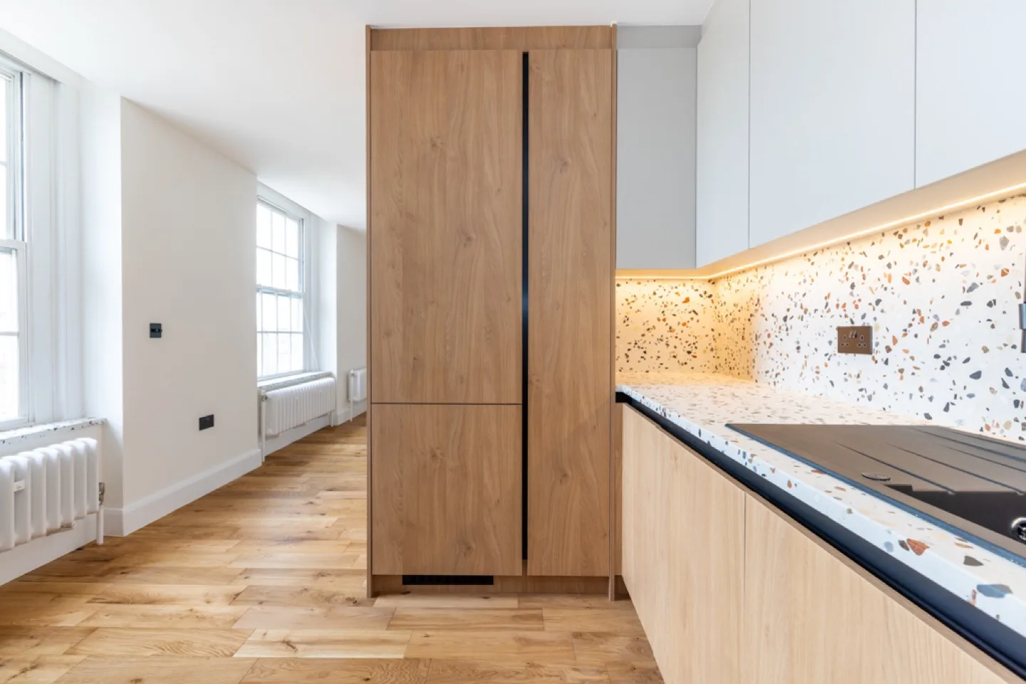 Tall oak larder unit housing the fridge, set against off-white wall units and terrazzo worktop