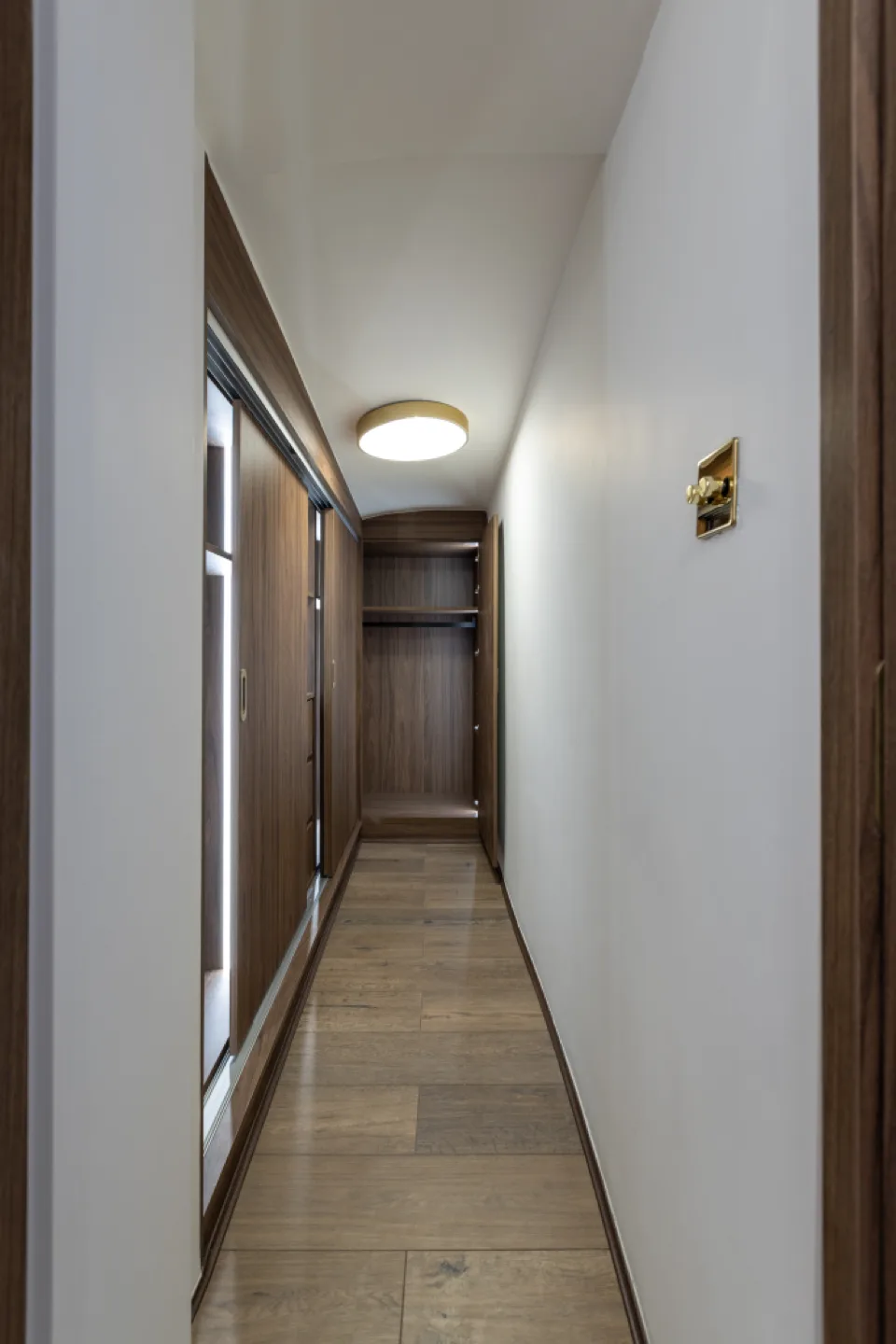 Narrow walk-in wardrobe lined with walnut units and sliding doors, with an open hanging rail at the far end