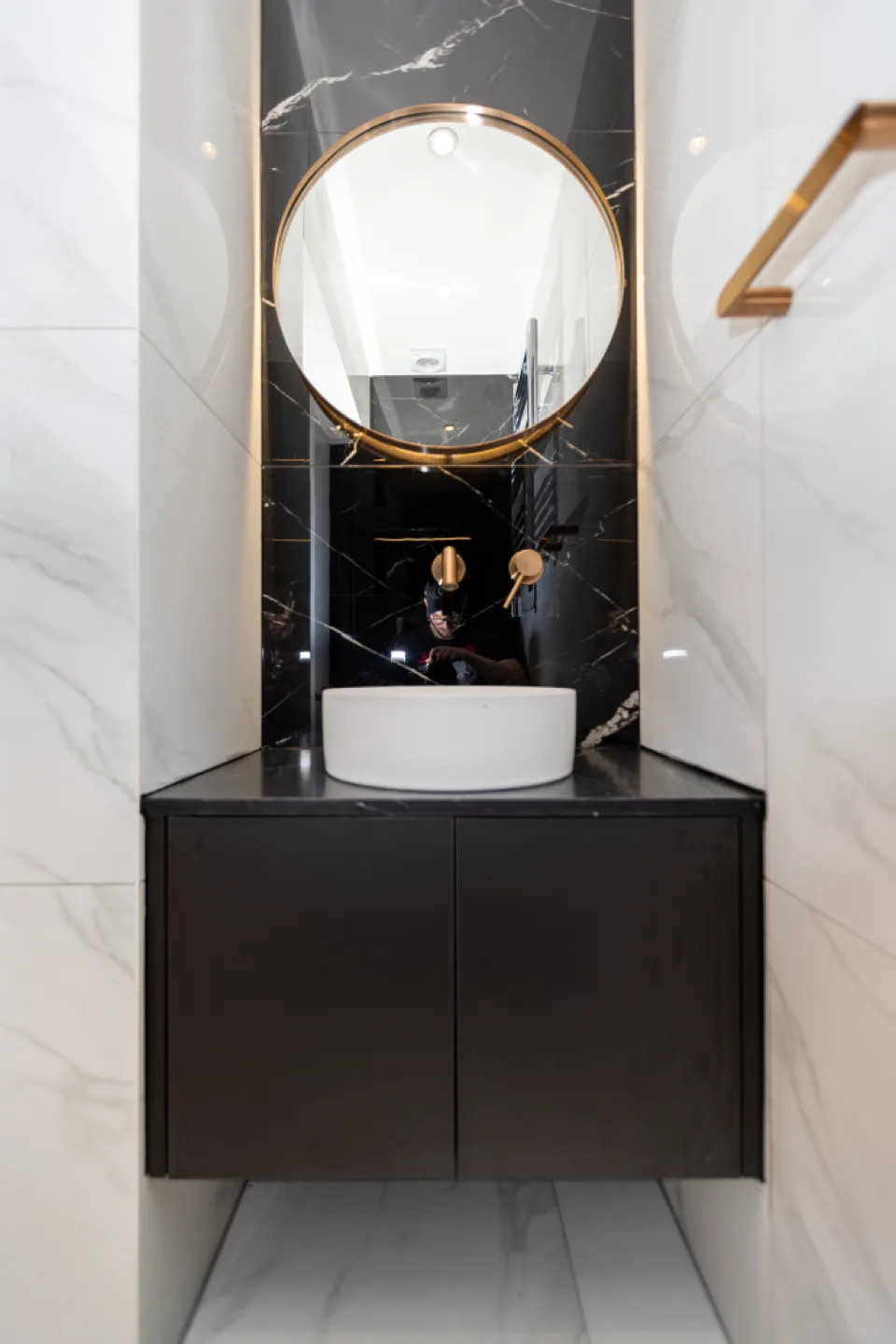 Matt black push-to-open bathroom vanity with white basin, round brass mirror and black marble feature wall