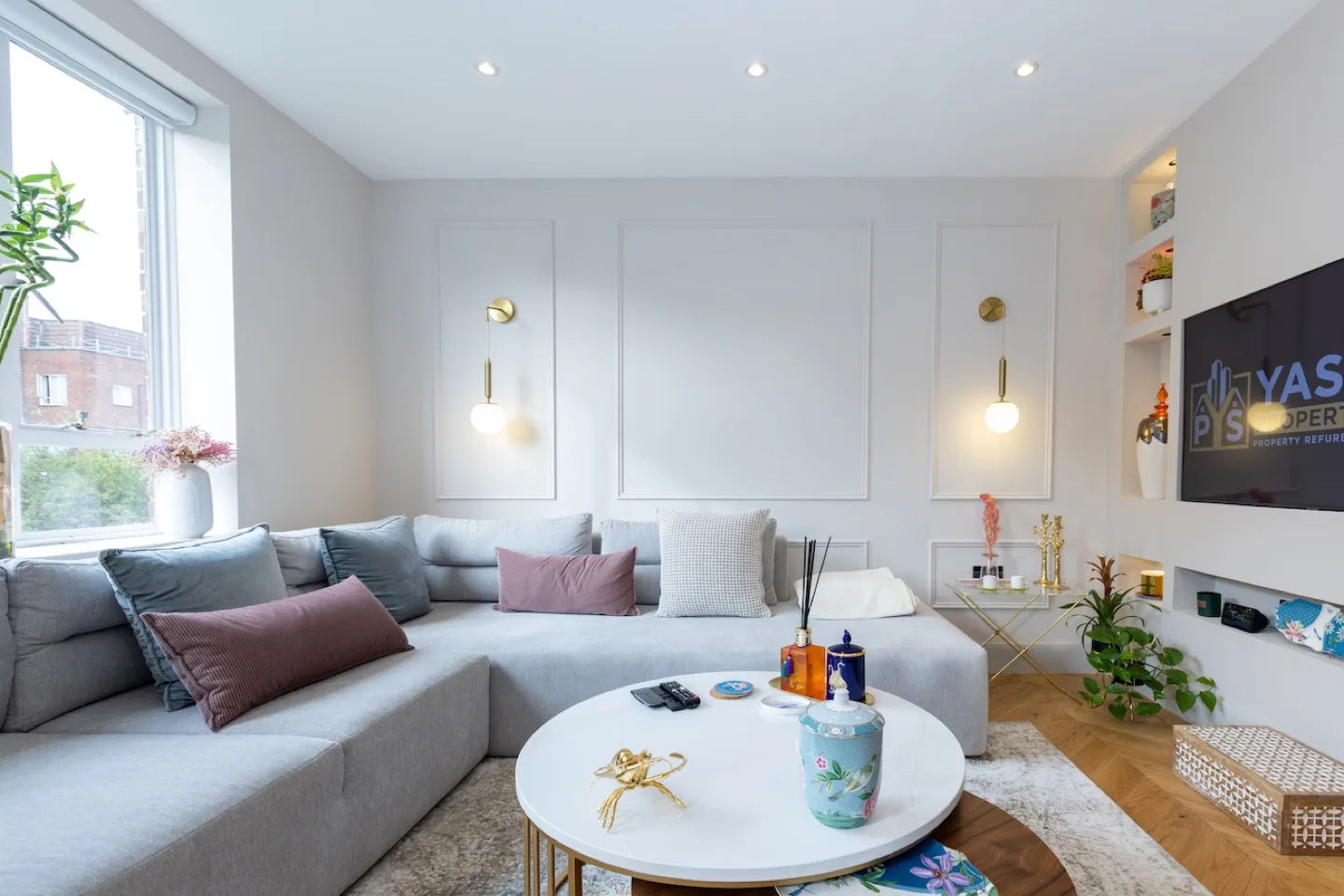 Living room with white wall panelling, grey corner sofa, brass wall lights and a round coffee table