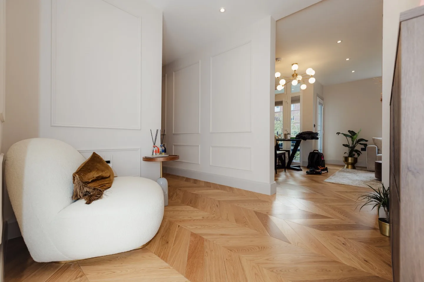 Herringbone oak flooring through the living area and hallway, framed by white panelling and a bouclé chair