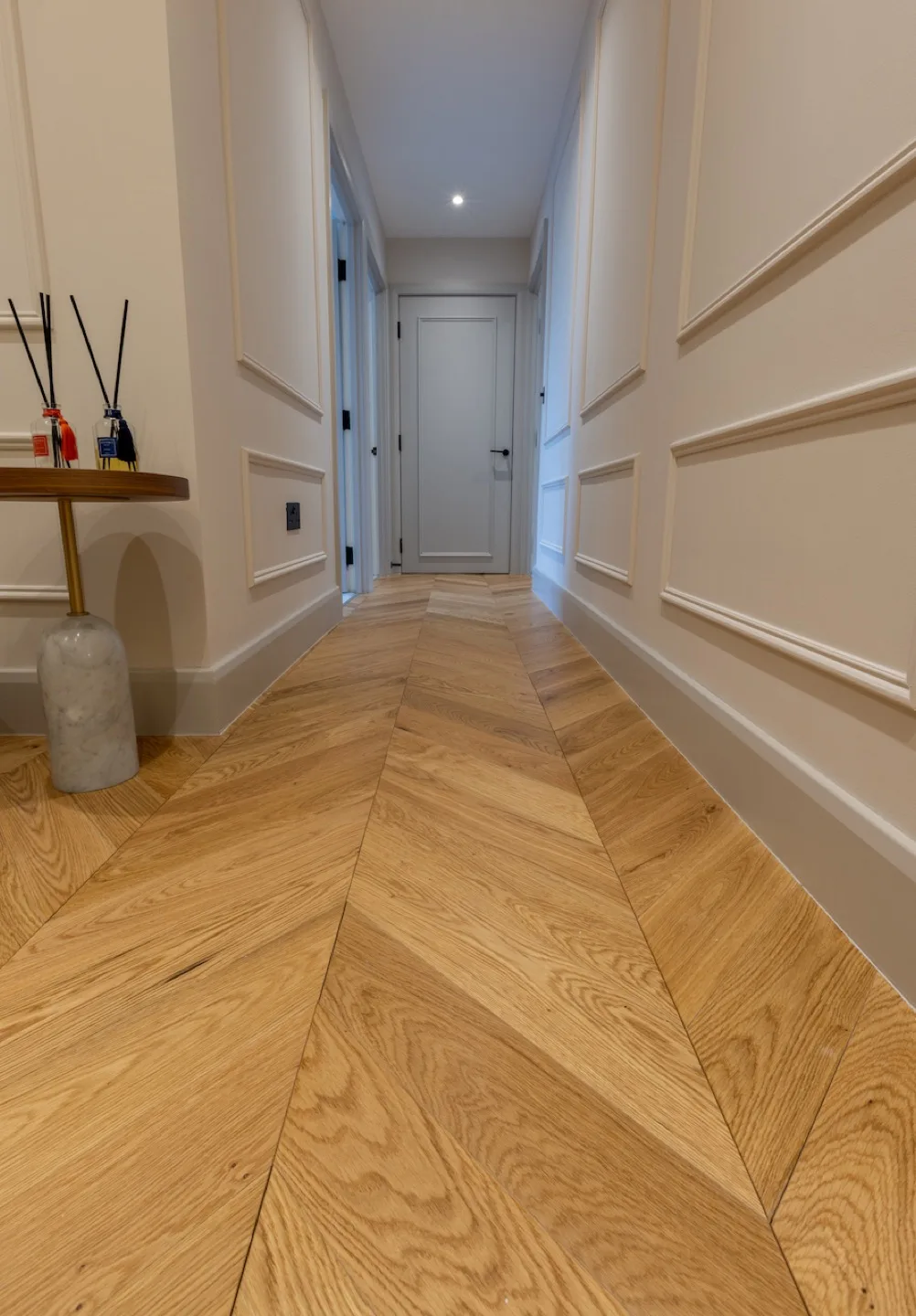 Chevron oak flooring running down the panelled foyer hallway towards a grey panelled door
